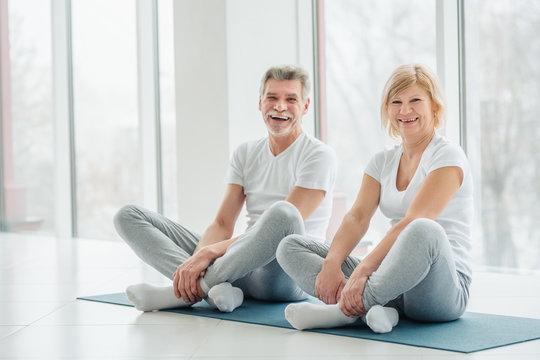 A beautiful senior family sits on a mat in a white fitness room before starting a sports lesson and laughs while looking at the camera. Sports lifestyle.