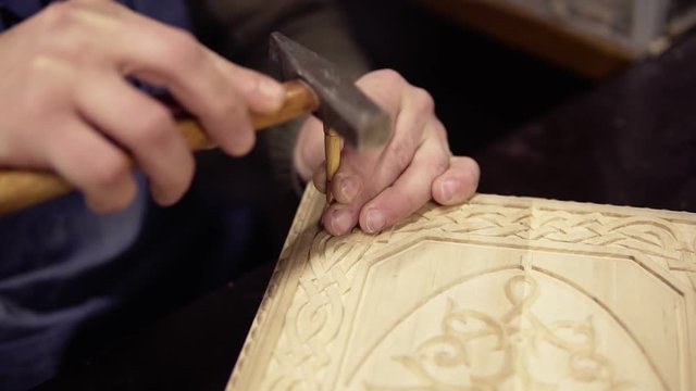 A craftsman carving wooden ornament on backgammon sitting in his working place. Close up footage of a man working on details using small wooden stick and hammer