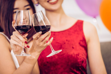 Two Asian women drinking red wine hand clink glasses together in party.