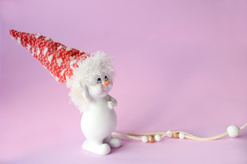 Funny cute snowman in a Christmas red hat next to a white ribbon and balls on a pink background...