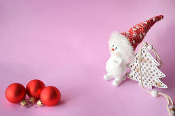 Funny cute snowman in a red hat near the Christmas tree and three red Christmas balls on a pink background with copy space. Christmas and New Year concept, minimalism.