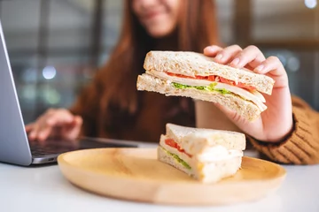 Abwaschbare Fototapete Snack Eine asiatische Frau, die Vollkornbrot hält und isst, während sie an einem Laptop arbeitet