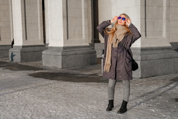 A beautiful woman in a gray coat and sunglasses walks on the street in the city.