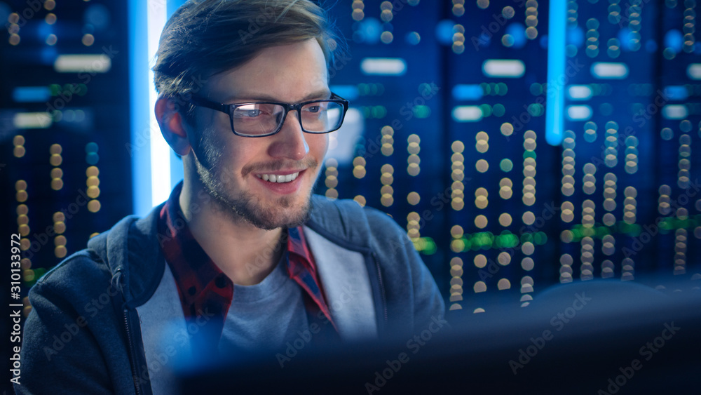 Poster Portrait of Smart SmilingFocused Young Man Wearing Glasses Working on Desktop Computer. In Background Technical Department Office with Functional Data Server Racks.