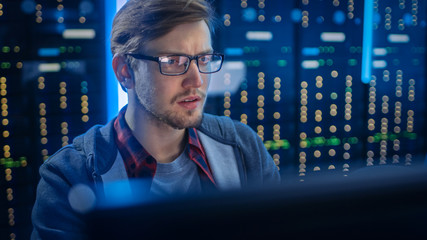 Portrait of a Smart Focused Young Man Wearing Glasses Сoncentrated on a Desktop Computer. In the Background Technical Department Office with Functional Data Server Racks.