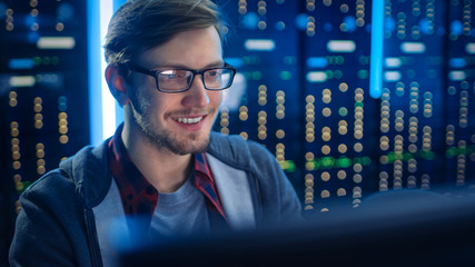 Portrait of Smart SmilingFocused Young Man Wearing Glasses Working on Desktop Computer. In Background Technical Department Office with Functional Data Server Racks.