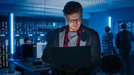 Fototapeta premium Portrait of a Smart Focused Young Man Wearing Glasses Holds Laptop. In the Background Technical Department Office with Specialists Working and Functional Data Server Racks