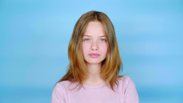 Happy teen girl in a pink pullover is laughing and looking at the camera. Blue background with copy space. Teenager emotions. 4k footage