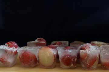 On a wooden Board and a black background are ice cubes with Isabella grapes.