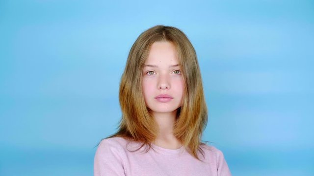 Teen girl in a pink pullover is posing and looking at the camera. Blue background with copy space. Teenager emotions. 4k footage