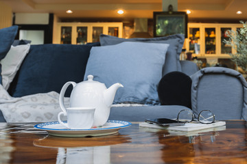 White teapot with cup and eyeglasses with cell phone on wooden table at home.