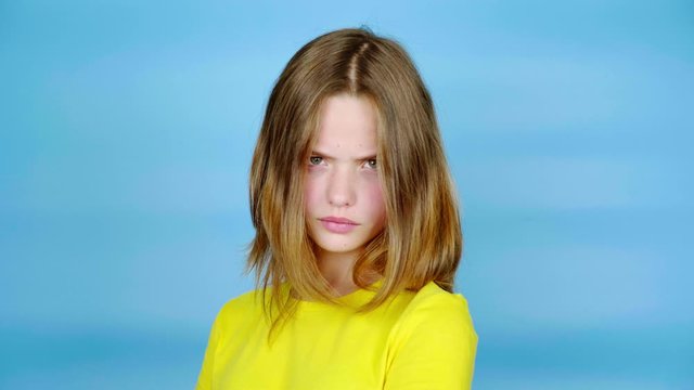 Teen girl in a yellow t-shirt is angry and looking at the camera. Blue background with copy space. Teenager emotions. 4k footage