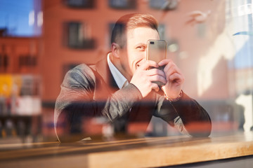 Happy male with phone in hand sitting in cafe, reflection of building in glass
