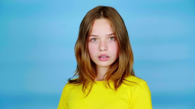 Teen girl in a yellow t-shirt is surprised and looks at the camera. Blue background with copy space. Teenager emotions. 4k footage