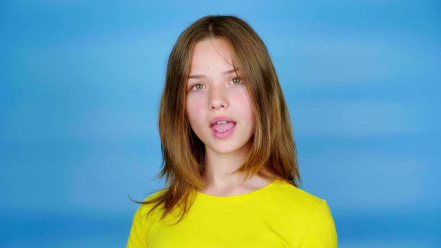 Teen girl in a yellow t-shirt is looking to the camera, nods her head and says yes. Blue background with copy space. Teenager emotions. 4k footage