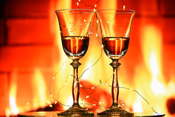 Two glasses of sparkling champagne wine on mustard fire in the fireplace. A cozy atmosphere of winter evenings by the fireplace, celebration of winter holidays.