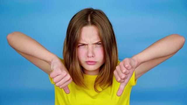 Angry teen girl in a yellow t-shirt is showing thumbs down, dislike and looking at camera. Blue background with copy space. Teenager emotions. 4k footage