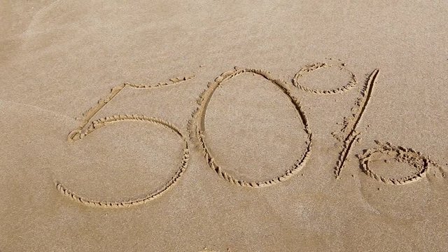 Handwritten inscription 50% on a sandy sea beach is washed by the sea wave with foam on a summer sunny day. Disappearing statistics and percent writing