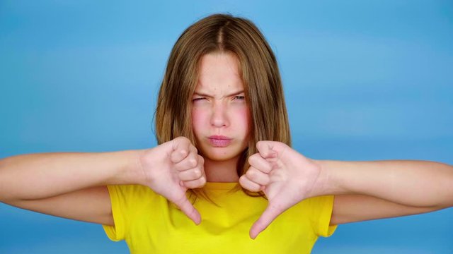 Angry teen girl in a yellow t-shirt is showing thumbs down, dislike and looking at camera. Blue background with copy space. Teenager emotions. 4k footage