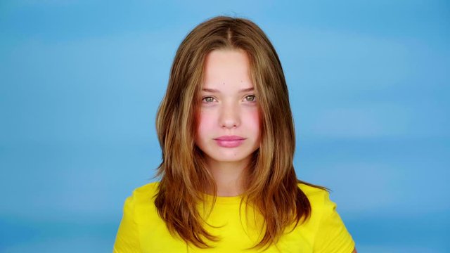 Teen girl in a yellow t-shirt is screaming and looking at camera, grimaces. Blue background with copy space. Teenager emotions. 4k footage