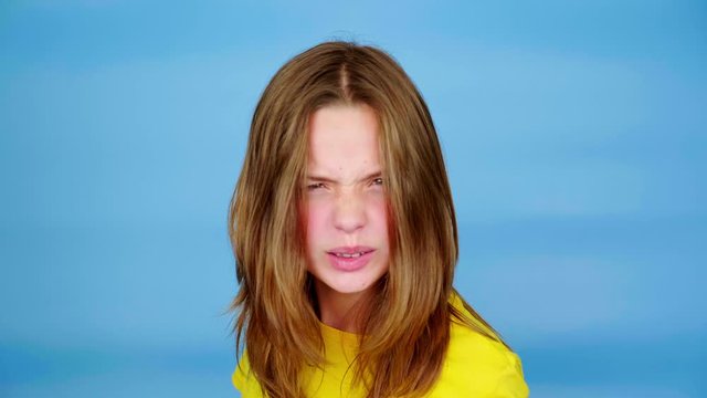 Angry teen girl in a yellow t-shirt is screaming and looking at camera, grimaces. Blue background with copy space. Teenager emotions. 4k footage