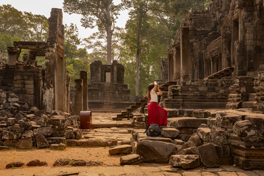 young female tourist in red dress taking a picture of the historic Bayon Temple, Angkor Wat, Cambodia