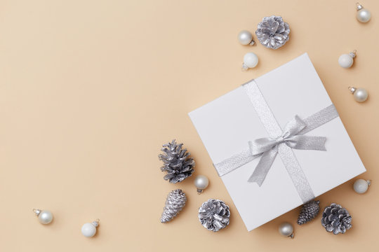 Flat Lay Composition Of A White Gift Box With A Silver Bow Surrounded With Silver And White Decorations On The Biege Background. Christmas Concept