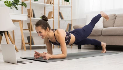 Muurstickers Fit vrouw doet yoga plank en kijkt naar online tutorials op laptop © Prostock-studio
