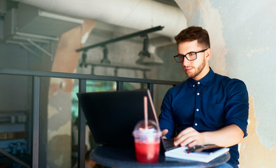 Young attractive hipster freelancer working remotely on laptop in loft cafe. Businessman browsing internet at coffeshop. Blogger posts to social media on travel using 5g internet. Telecommuting theme.
