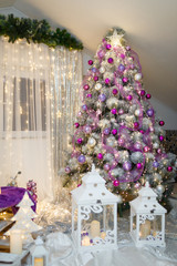 Christmas or New Year tree decorated with purple baubles, shiny star on top and garland lights with two white lanterns on the foreground. Silver curtains on the window on background.