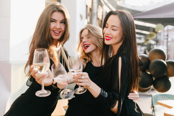 Portrait of three sisters in black outfit celebrating important event with champagne. Inspired long-haired woman looking away while clink glasses with friends in city background.