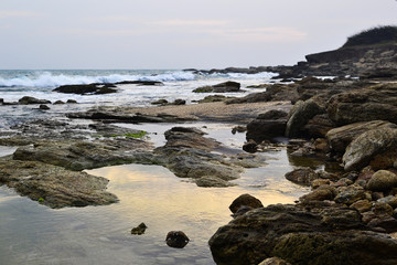 Fototapeta na wymiar Big rocks on the beach. Waves in the ocean. Sunset sky reflected in water. Rocky coastline in Kanyakumari, Tamil Nadu, India.