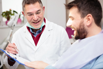 Smiling middle aged male dentist consulting with young patient, filling in questionnaire and explaining therapy procedure. Healthcare and medicine concept.