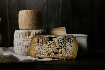 Different homemade cheeses on a dark wooden background. Pieces and cheese heads of Gorgonzola, Asiago, Maasdam, Gauda, Paramezan and Edam. Goat and cow cheese on baking paper. Handwork. Close up