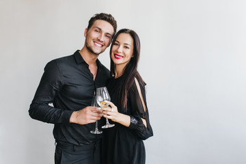 Stunning brunette woman clink glasses with handsome husband during friend's party. Indoor portrait of blissful man enjoying event with girlfriend in black dress, smiling on white background.