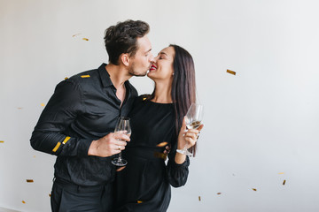 Black-haired man in stylish shirt kissing his wife on light background. Cute couple with glasses of wine celebrating anniversary under sparkle confetti.