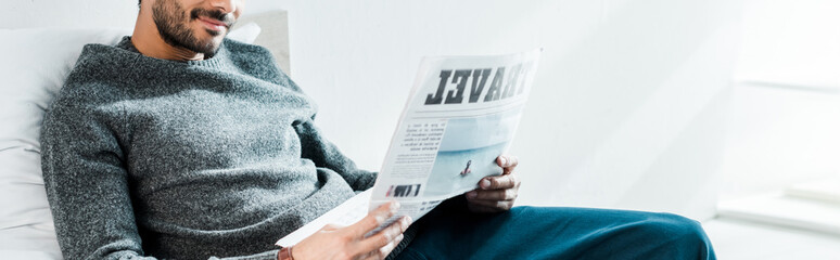 panoramic shot of smiling bi-racial man reading newspaper with lettering travel