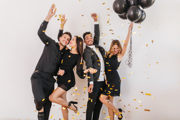 Graceful brunette girl with pale skin throwing sparkle confetti at birthday party. Good-looking curly woman with helium balloons standing on one leg while posing for photo.