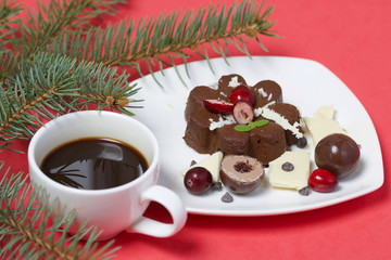Chocolate fudge garnished with chocolate, mint and cranberries. Lies on a white plate, on a white background. Nearby a cup of coffee and a branch of spruce.