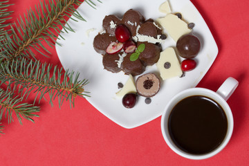 Chocolate fudge garnished with chocolate, mint and cranberries. Lies on a white plate, on a white background. Nearby a cup of coffee and a branch of spruce.