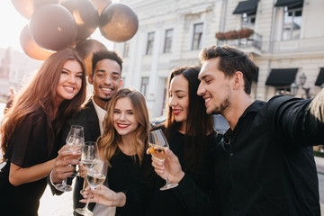 Blissful african man holding bunch of balloons and glass of wine in front of old building. Outdoor portrait of excited friends enjoying champagne during meeting after workday.