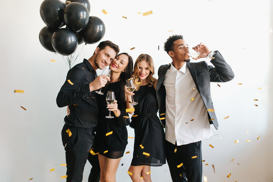 Indoor portrait of romantic pale girl with shiny curls enjoying wine with friends. Ladies in black dresses having fun at party with guys and dancing under confetti.