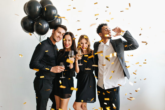 Happy couple with balloons celebrating anniversary with their friends. Indoor portrait of students in beautiful attires having fun at event with confetti and drinking wine.
