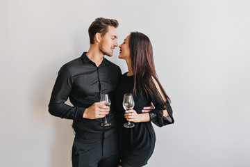 Married couple kissing at party, holding glasses of champagne in hands. Indoor portrait of confident man in black clothes looking with love at girlfriend during event.