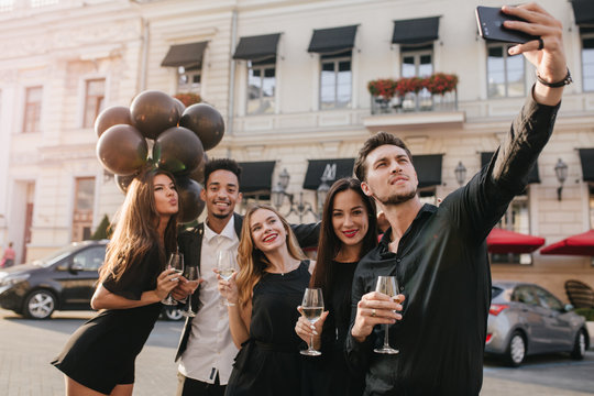 Brunette girl standing beside african guy with balloons and posing with kissing face expression. Glad man in trendy black attire making selfie during outdoor party with friends.