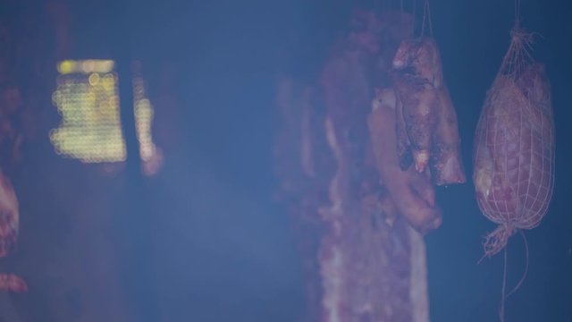 Ham and bacon hanging and drying in smokehouse on traditional way on blue smoke.  Slow motion.  The sunrays pierce through the blue smoke.