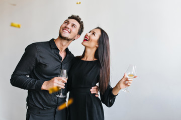 Portrait of excited young woman embracing with husband at event and posing with confetti. Happy dark-haired girl and smiling man with glass of champagne isolated on white background.