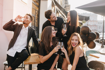 Brunette man in stylish outfit looking at his girlfriend with love while resting in city with friends. Outdoor portrait of glad african guy drinking champagne, posing in cafe with colleagues.