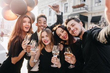 Portrait of long-haired brunette woman holds bunch of helium balloons and posing with surprised face epxression. Handsome man in black shirt making selfie with friends on city background.