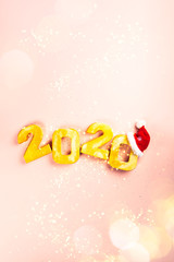 Naklejka premium Happy New Year 2020. Golden digits 2020 with christmas hat are on pink background with glitter. Holiday Party Decoration or postcard concept with top view and copy space.